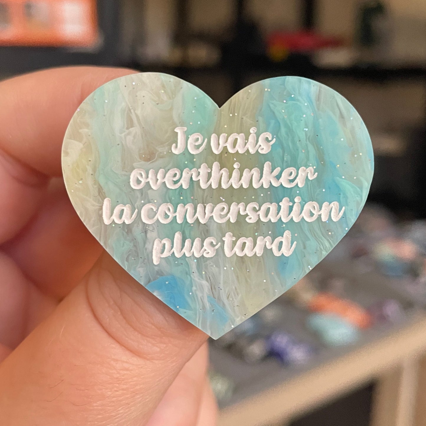 Broche "Je vais overthinker la conversation plus tard" en acrylique marbrée semi-transparente bleue, turquoise, blanche et jaune à paillettes