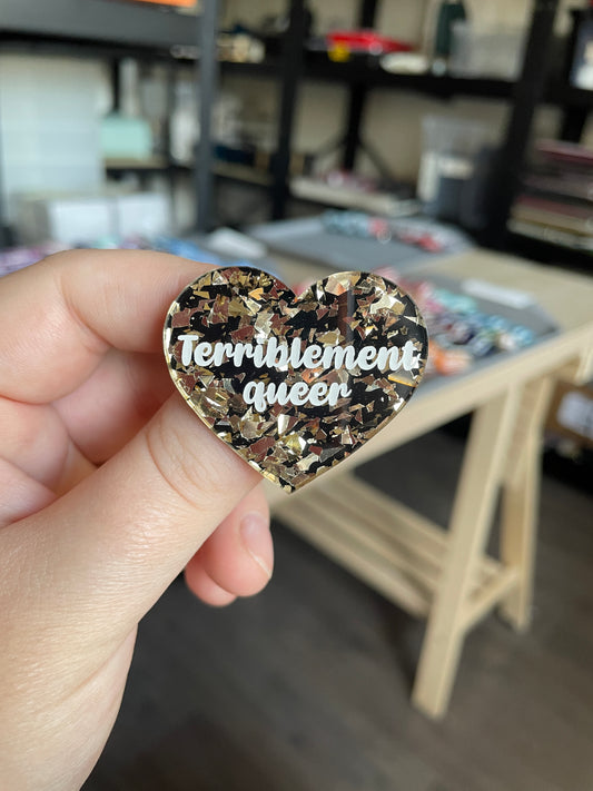 [CONTRÔLE TECHNIQUE] Broche "Terriblement queer" en acrylique noire avec des éclats de confettis dorés clairs