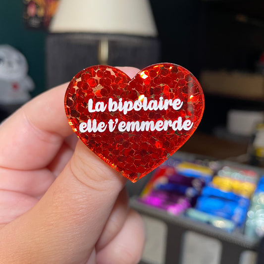 Broche "La bipolaire elle t'emmerde" en acrylique semi transparente avec des confettis rouges