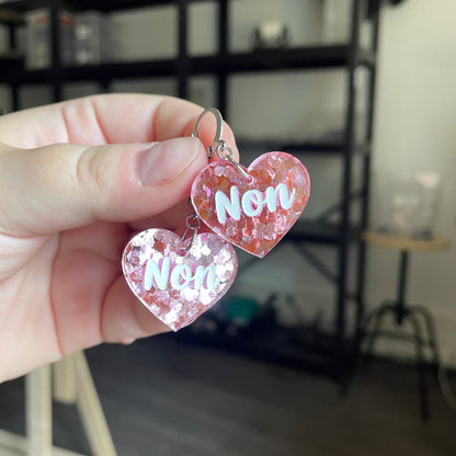 Petites boucles d'oreilles coeurs pendantes "Non" en acrylique semi transparente avec des confettis roses
